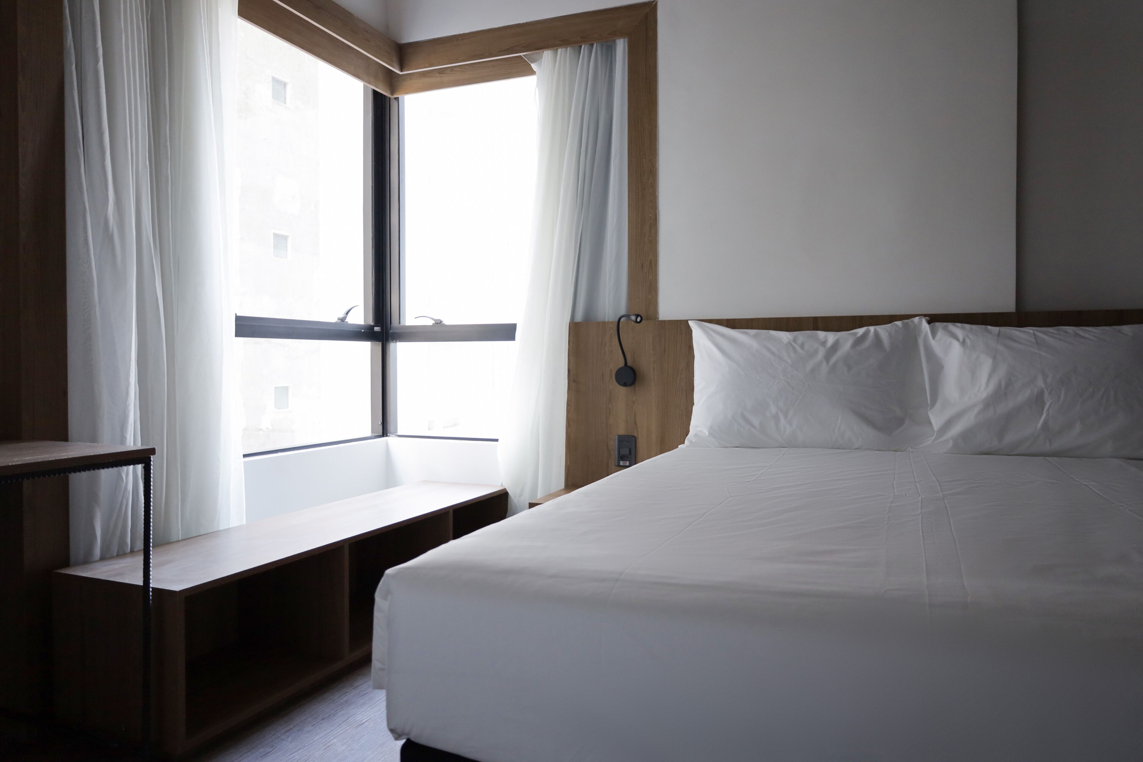 Quarto de hotel com uma cama grande, uma mesa de cabeceira de madeira, um banco e uma janela com cortinas.