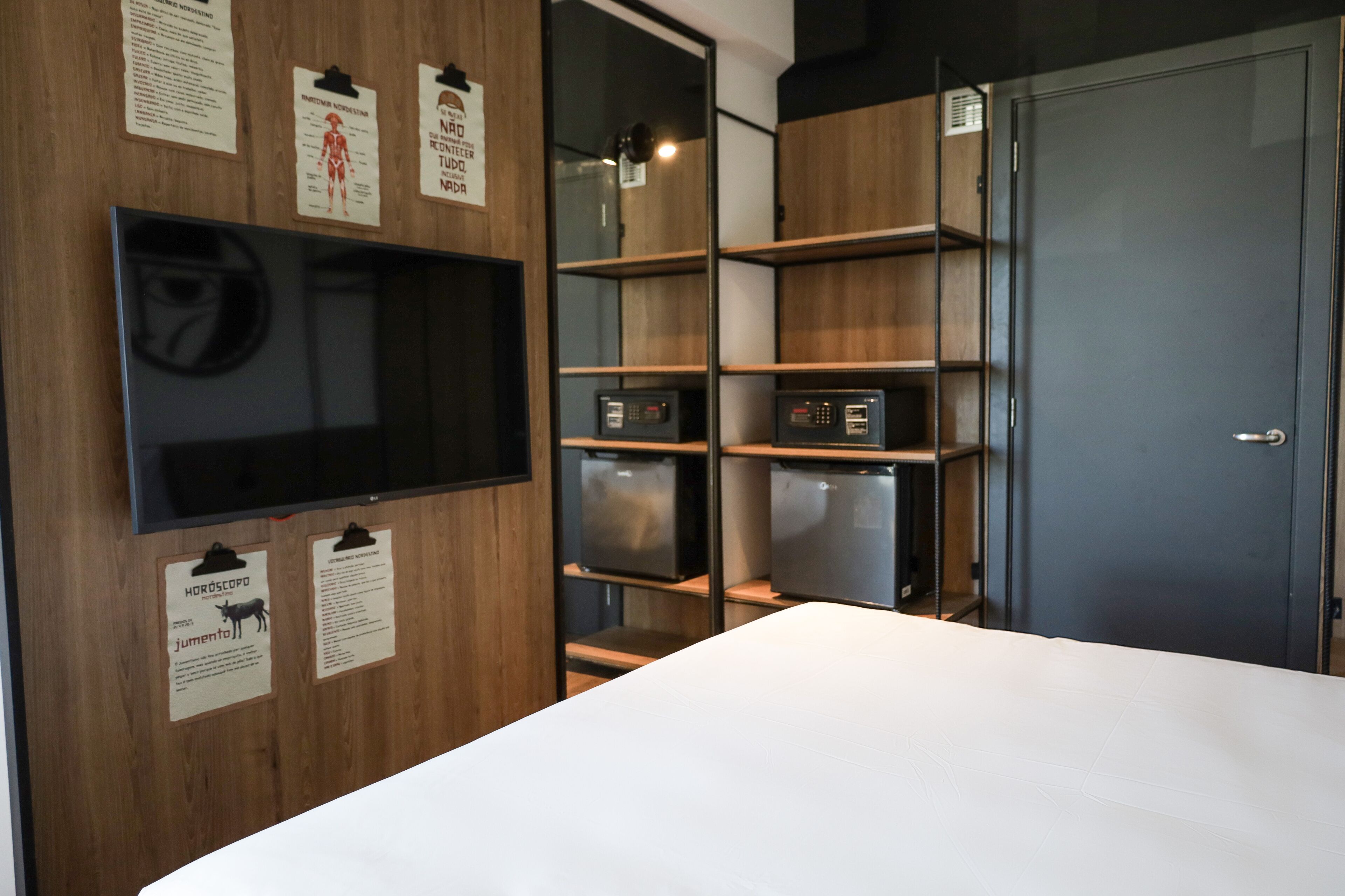 A hotel room with a large flat-screen TV mounted on the wall, wooden shelving units, and a door.