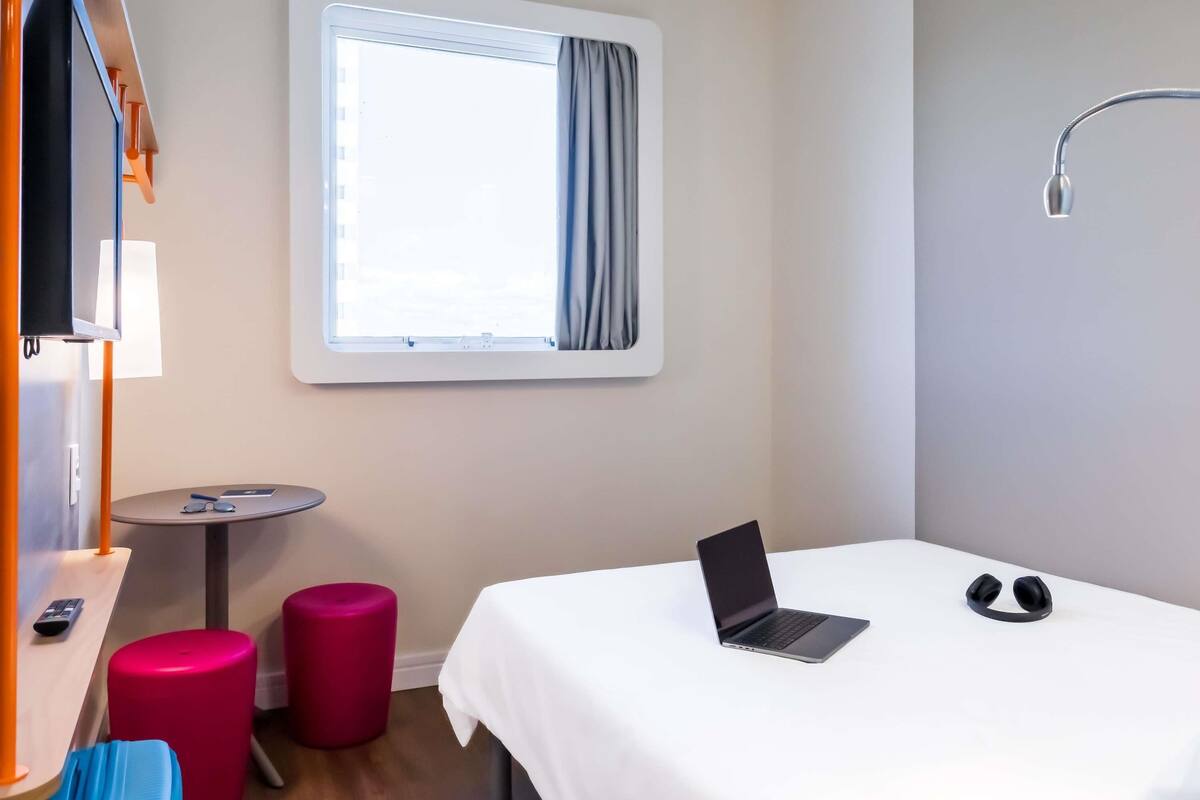A hotel room with a bed, a laptop, a pair of headphones, a small table with a lamp, and a window with curtains.