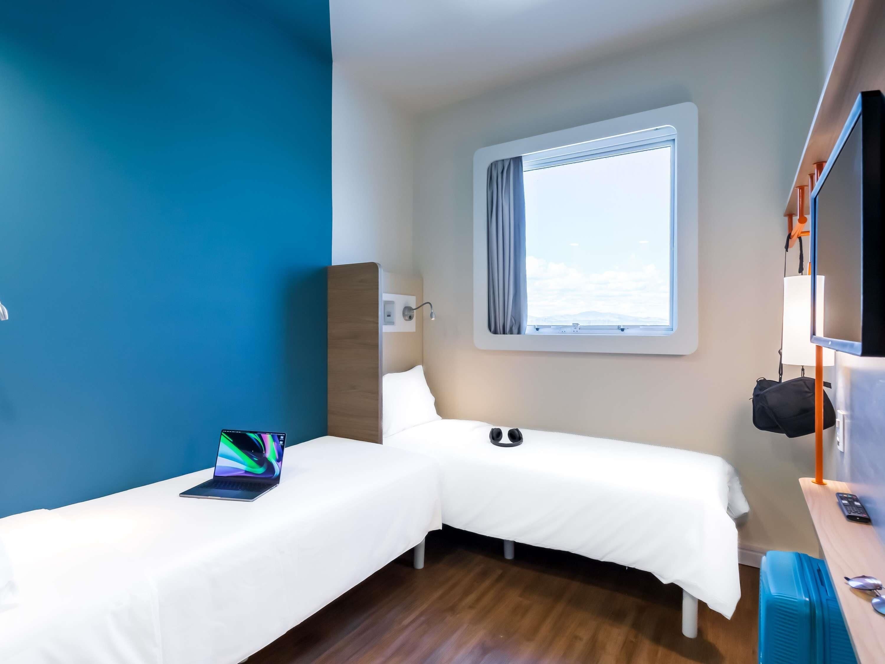 A hotel room with two single beds, a desk with a computer, a window with curtains, and a TV.