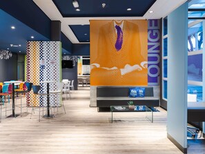A modern lounge area with a large orange wall featuring a white horse silhouette, a glass coffee table, and colorful chairs. - Ibis Budget Pindamonhangaba (Pindamonhangaba)