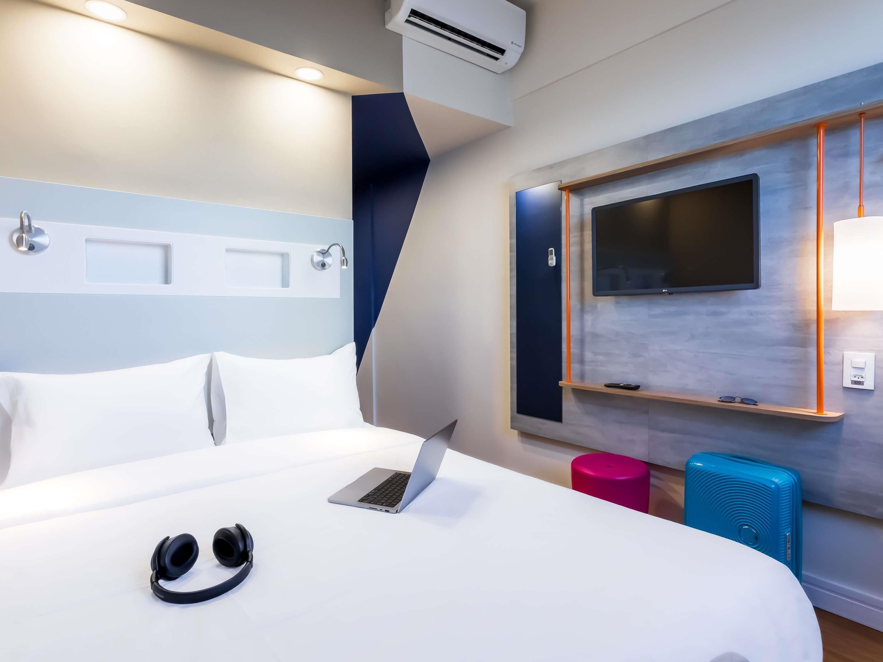 Une chambre d’hôtel équipée d’un lit, d’un ordinateur portable, d’écouteurs, d’une télévision et d’une petite table avec une télécommande.