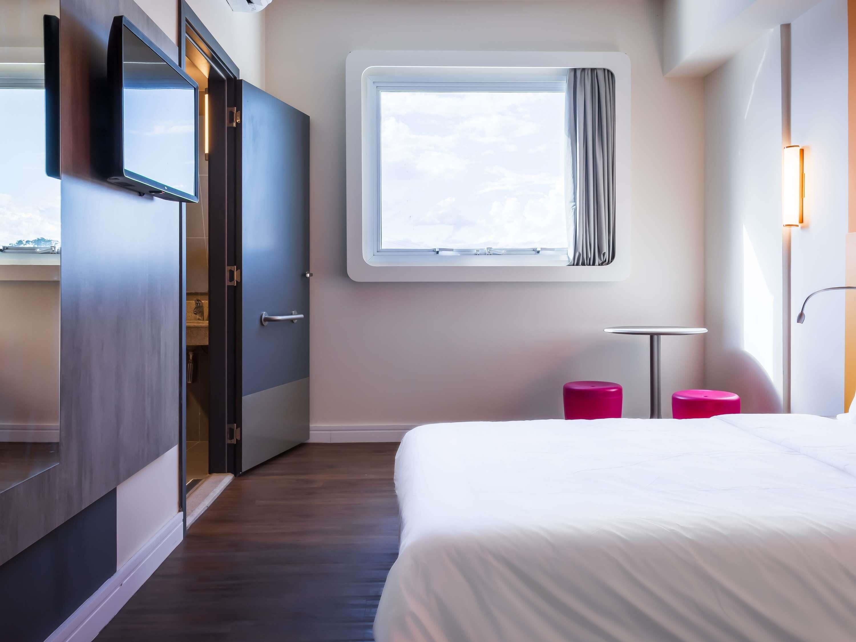 A hotel room with a bed, a TV mounted on the wall, a window with curtains, a small table with a lamp, and a pink stool.