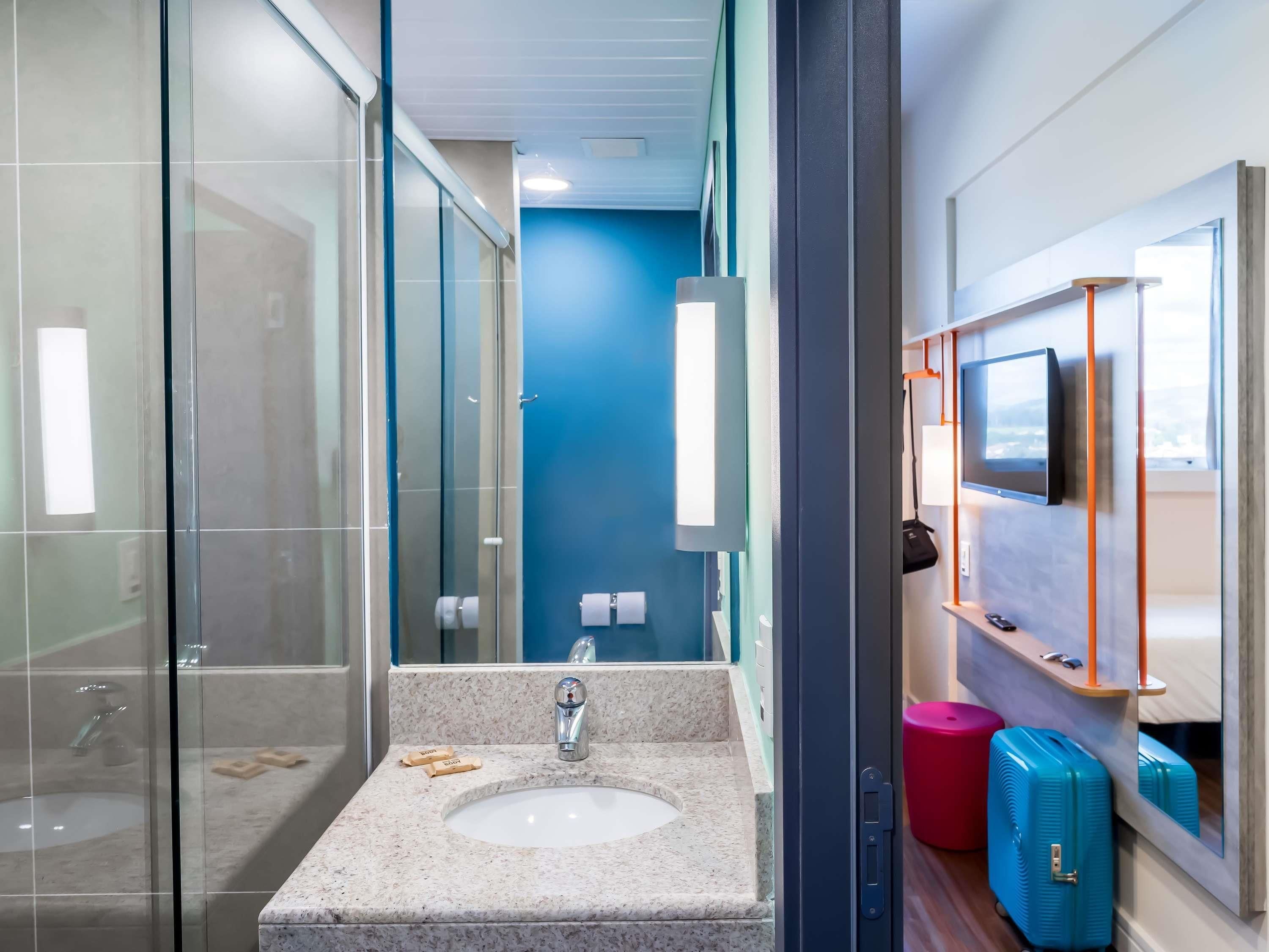 A modern bathroom with a glass shower enclosure, a sink with a marble countertop, a toilet, and a mirror with a mounted television.