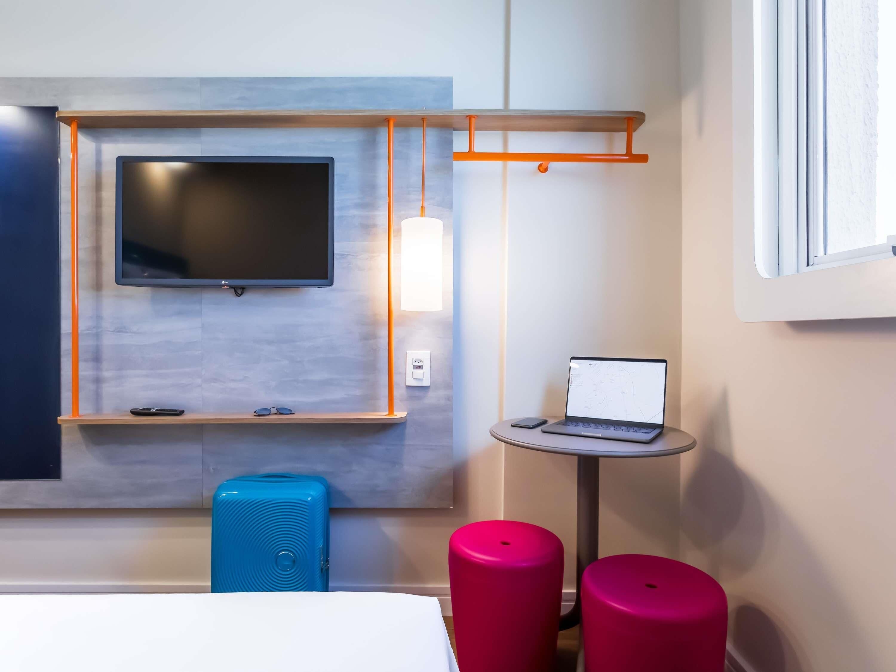 A modern hotel room with a flat-screen TV mounted on the wall, a small table with a laptop, a blue suitcase, and two red stools.
