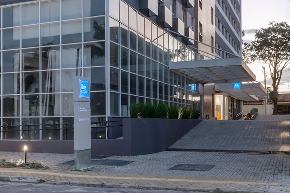 A modern hotel with a glass facade, a sign for ibis budget, and a landscaped entrance area. - Ibis Budget Pindamonhangaba (Pindamonhangaba)