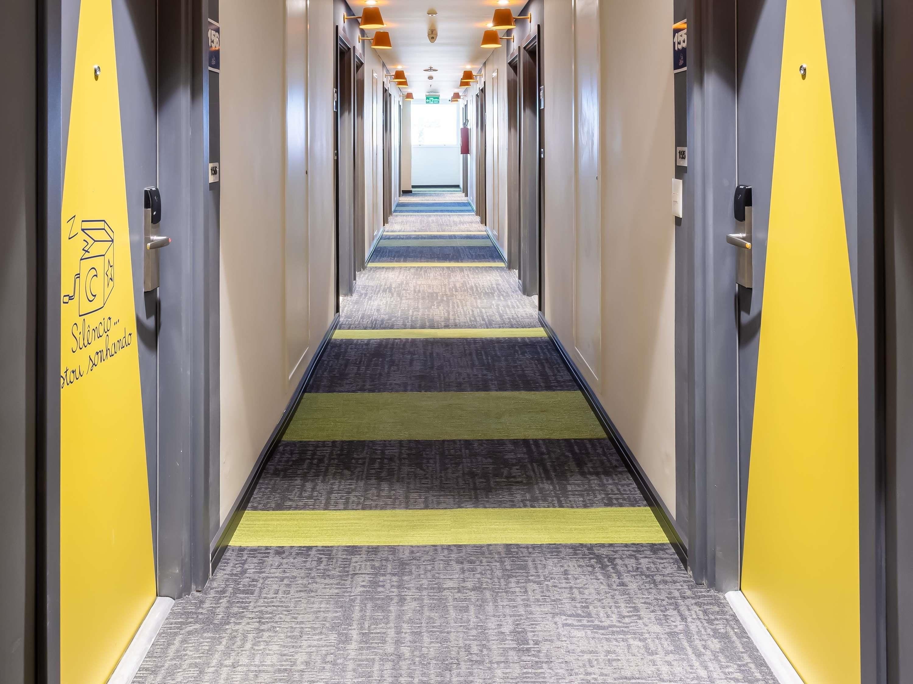 Un pasillo de hotel con puertas a ambos lados, una alfombra estampada y un cartel en la pared izquierda.