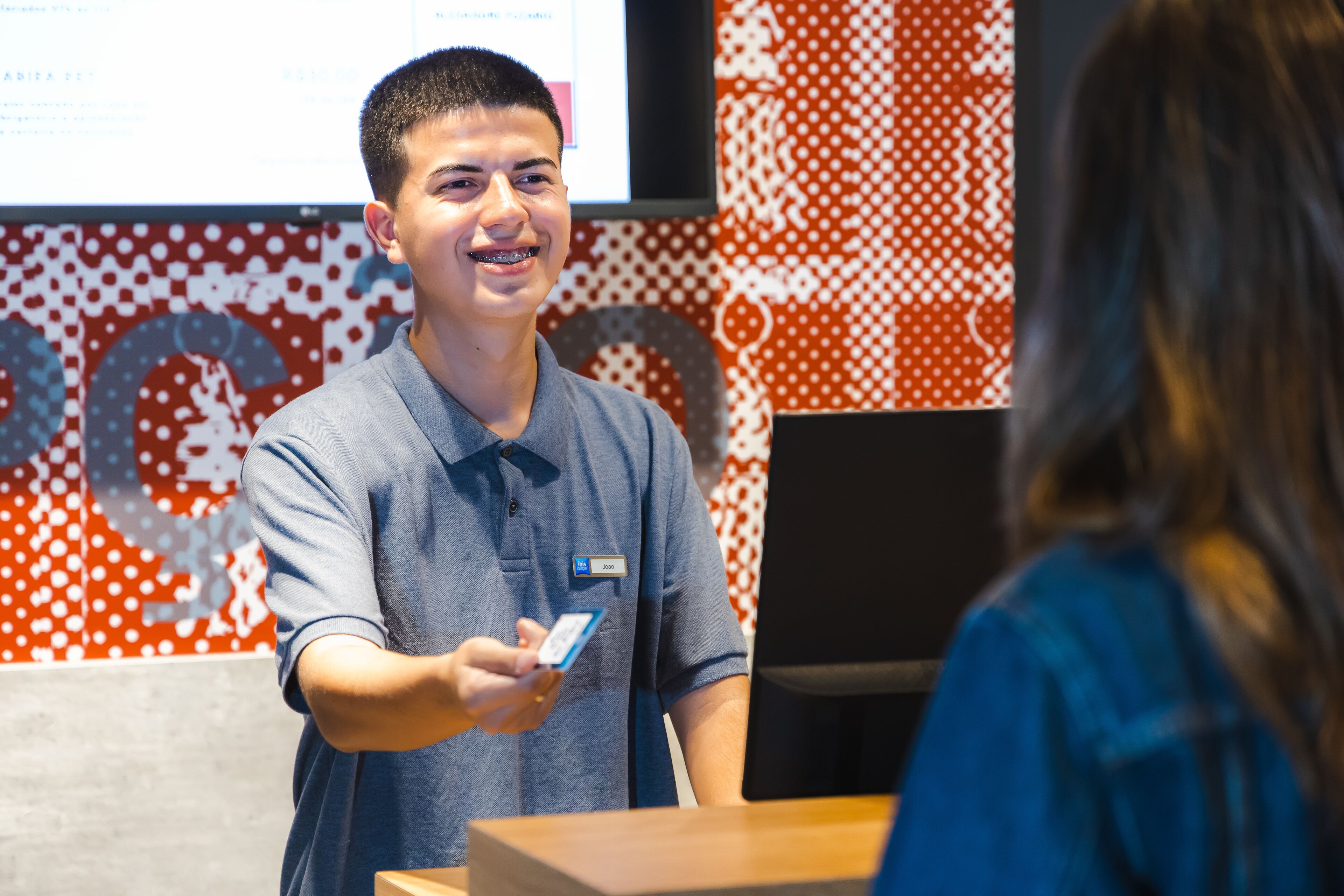 A person in a blue shirt holding a card, standing in front of a counter with a patterned wall in the background.