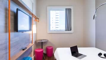 Habitación de hotel con cama, laptop, televisor montado en la pared, ventana con cortinas, mesita con lámpara y maleta.