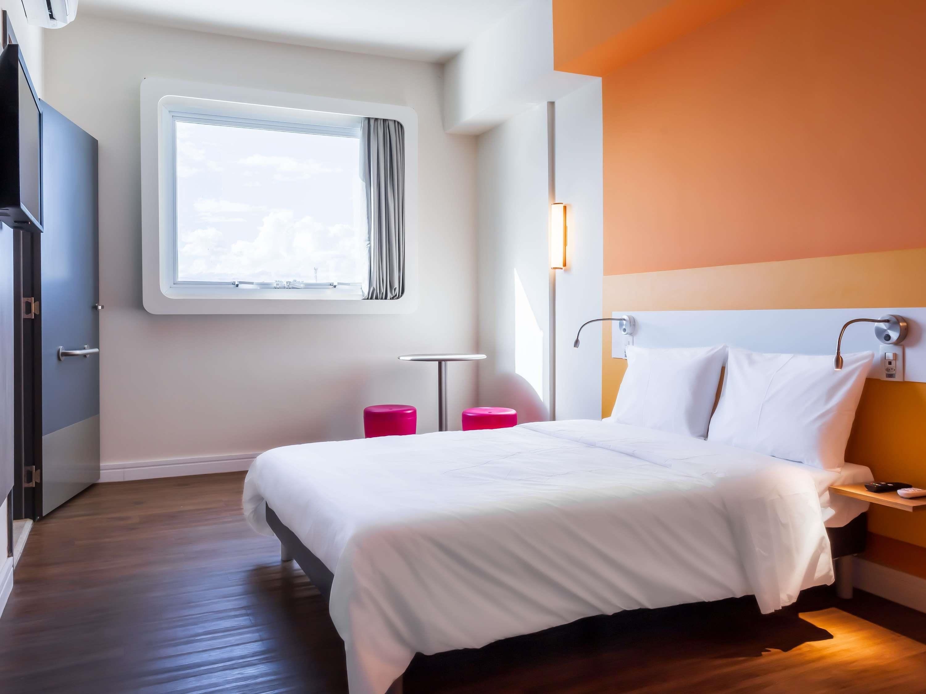 Una habitación de hotel con cama, mesita de noche, cortinas en la ventana, una lámpara de pared y vista al cielo.