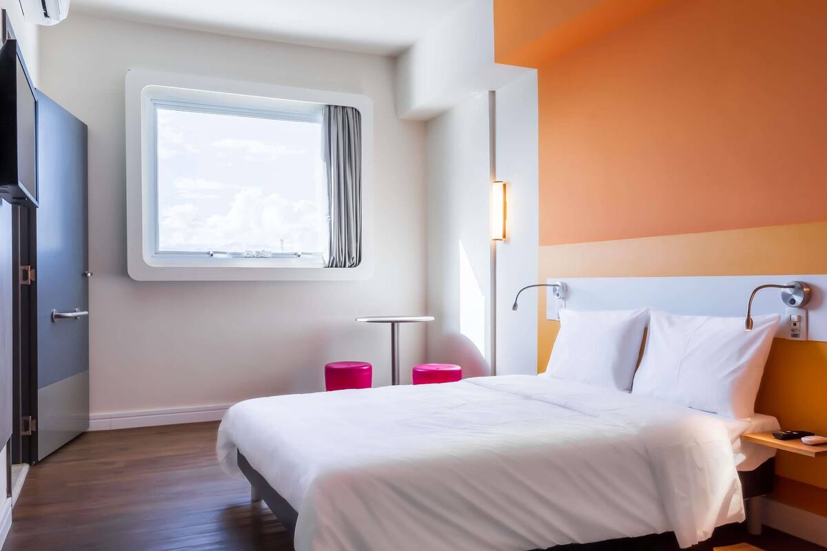 Una habitación de hotel con cama, mesita de noche, cortinas en la ventana, una lámpara de pared y vista al cielo.