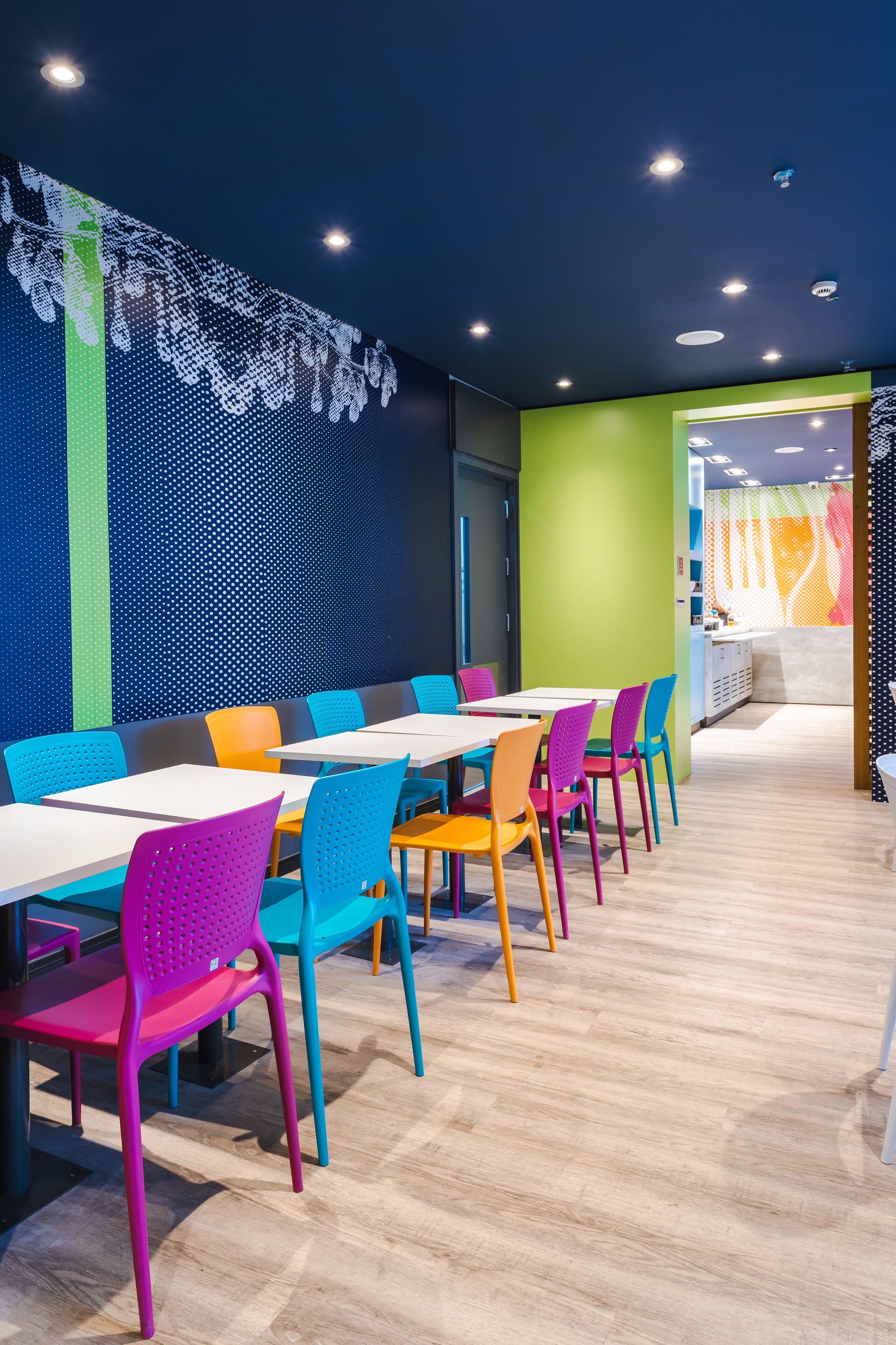 A brightly lit dining area with colorful chairs and tables, a patterned wall, and a view into another room.