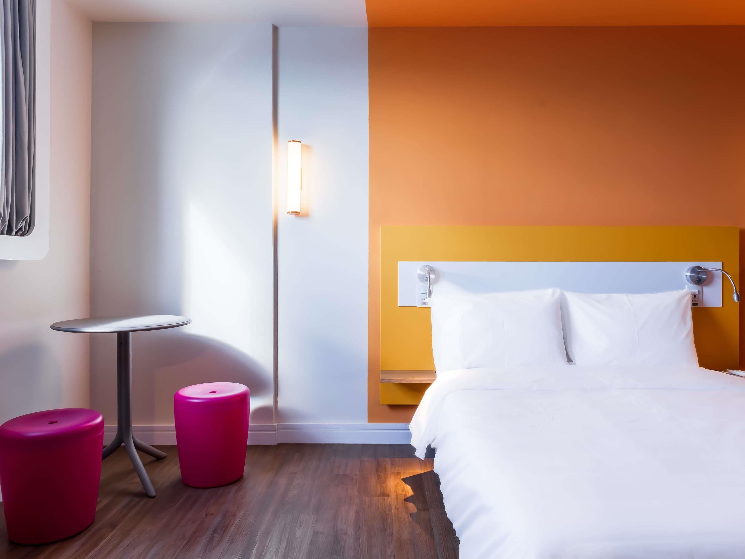 A hotel room with a bed, a small table, two pink stools, and a wall-mounted lamp.