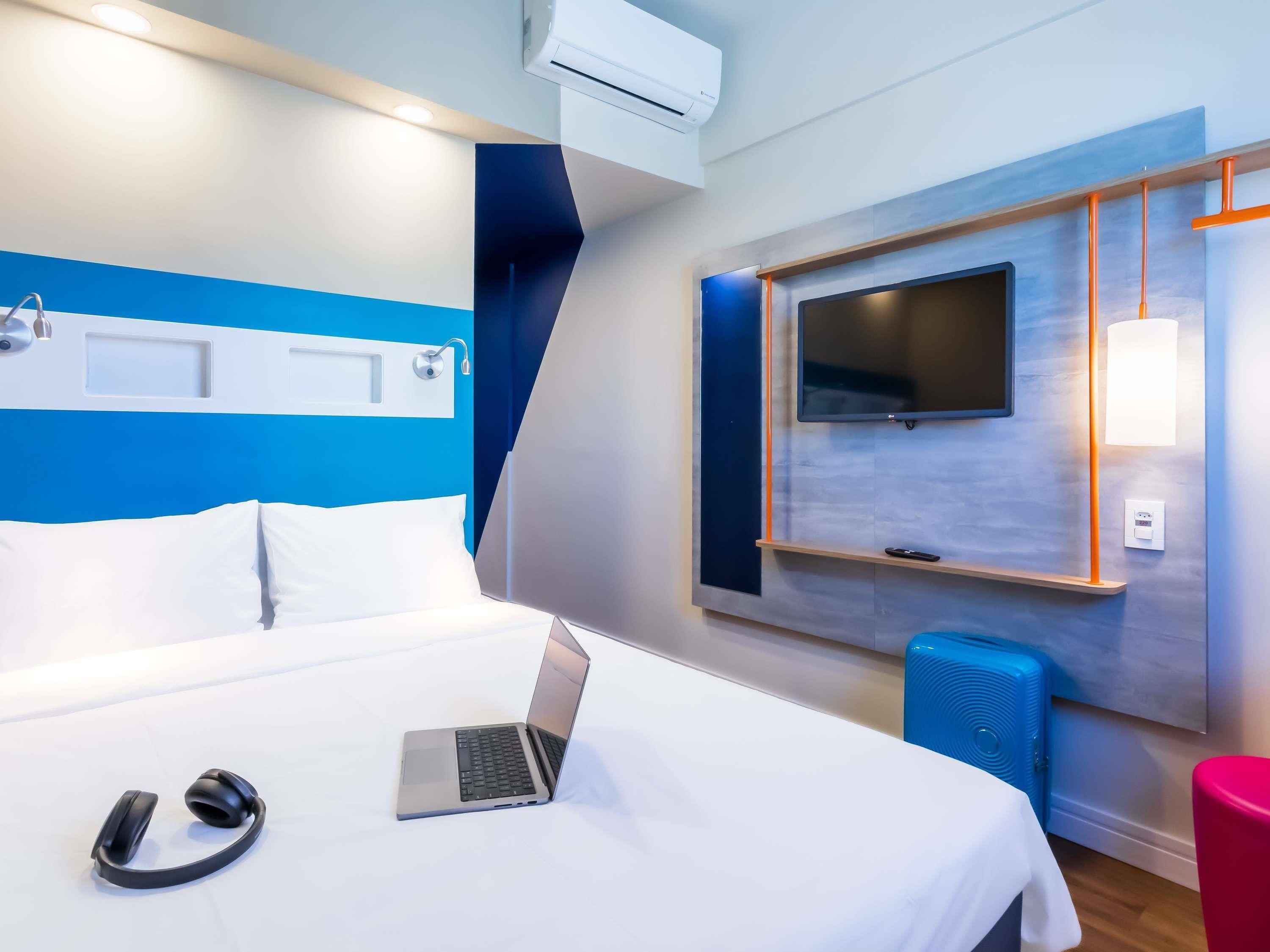 A modern hotel room with a bed, a laptop, headphones, a TV, and a blue speaker.