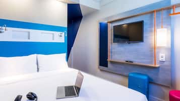 A modern hotel room with a bed, a laptop, headphones, a TV, and a blue speaker.