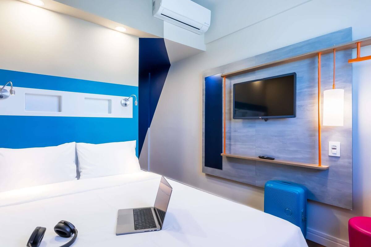 A modern hotel room with a bed, a laptop, headphones, a TV, and a blue speaker.