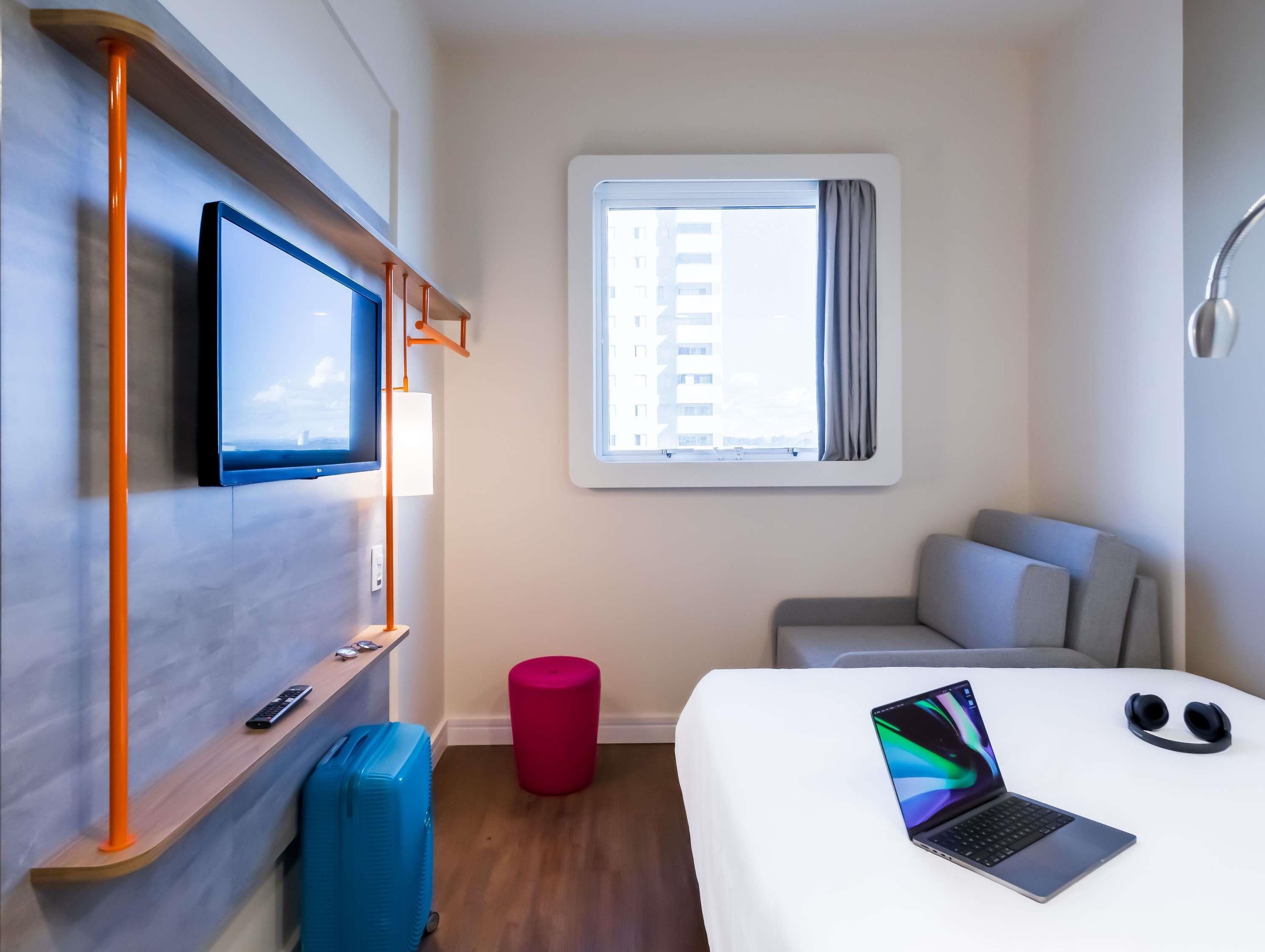 A hotel room with a bed, a desk with a laptop, a TV, a window with curtains, and a suitcase.
