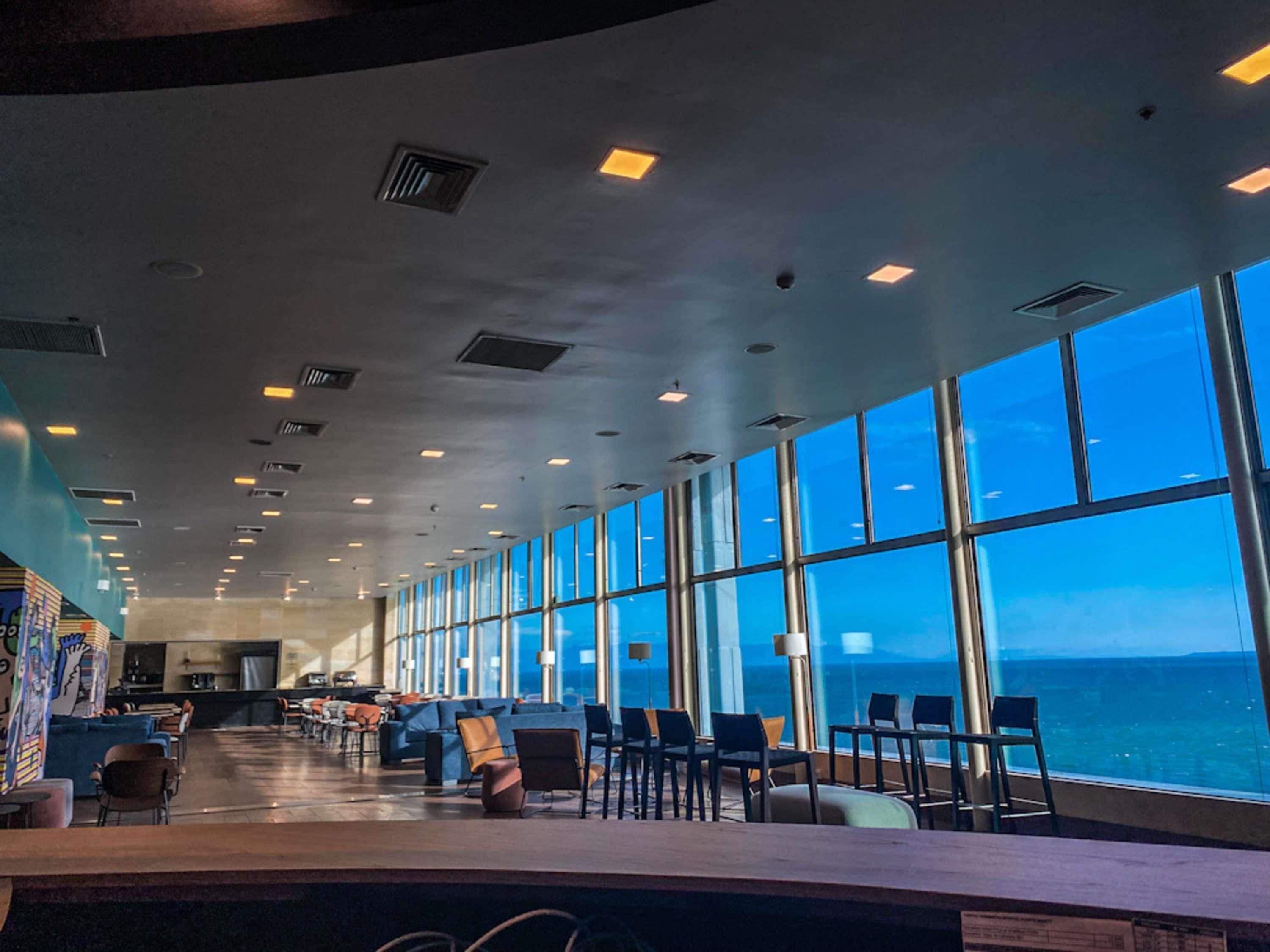 A spacious indoor area with a long row of tables and chairs, large windows offering a view of the ocean, and a high ceiling with recessed lighting.