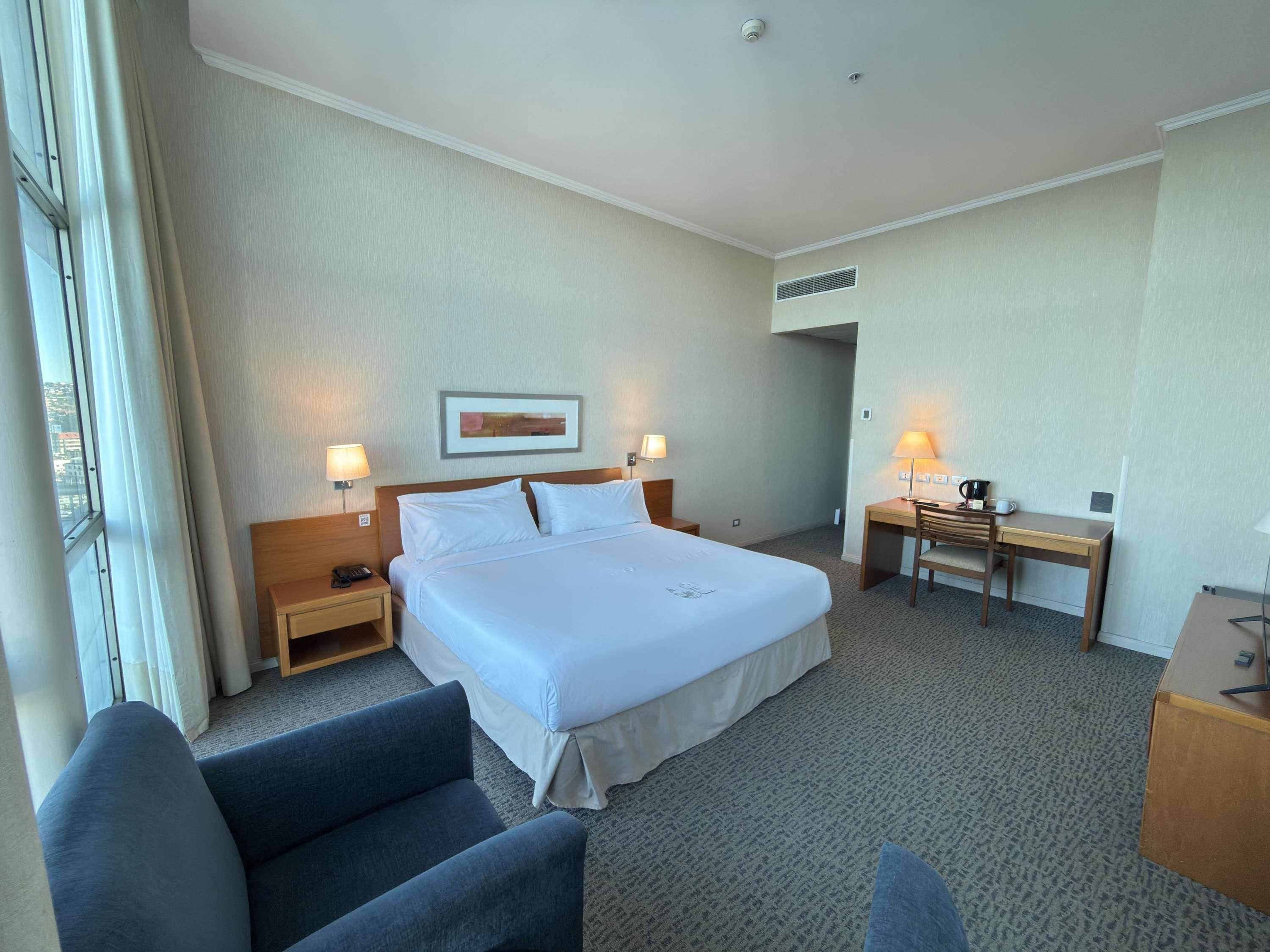 A hotel room with a large bed, two bedside tables with lamps, a desk with a chair, and a window with curtains.