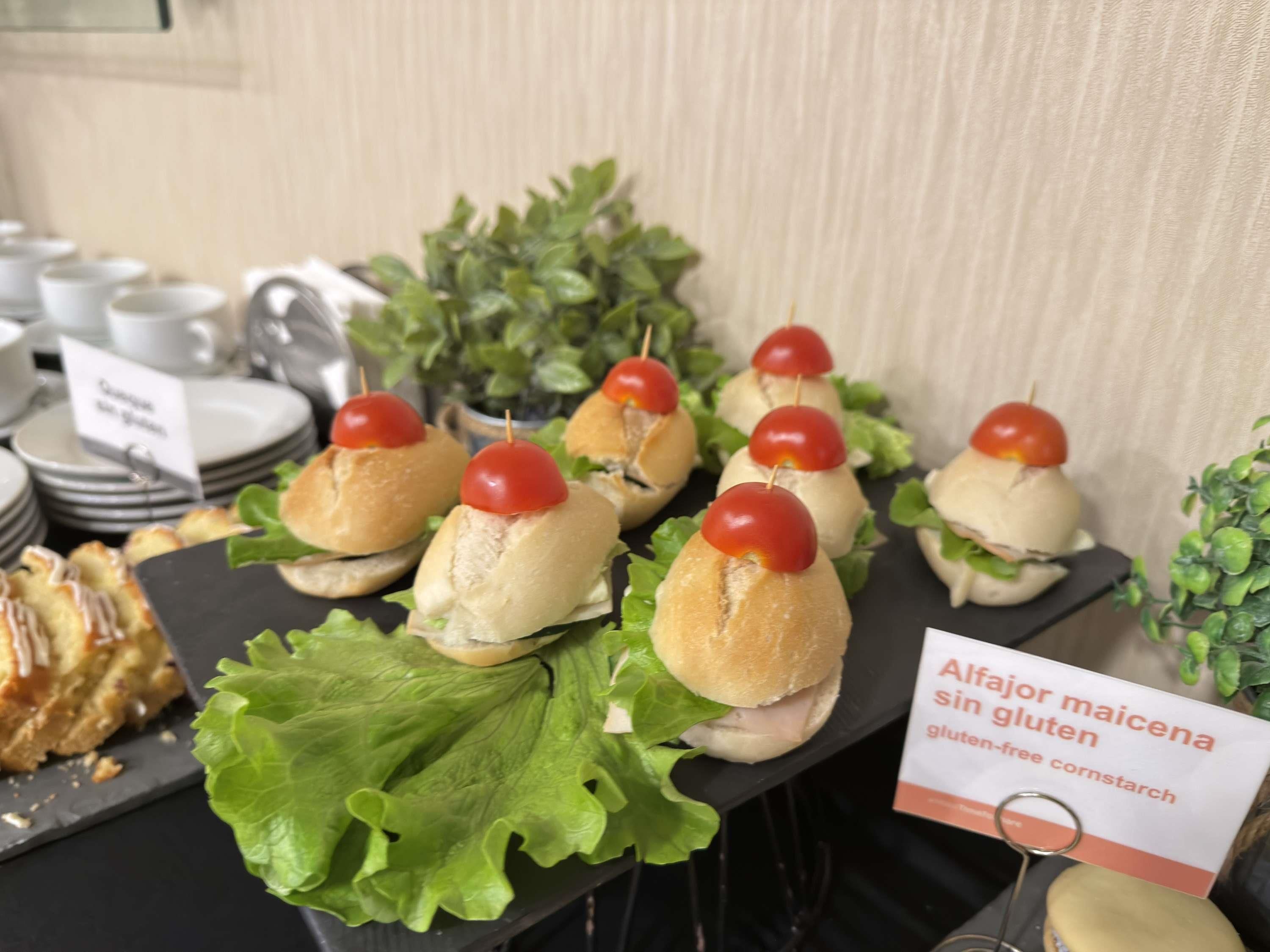 Una mesa de buffet con panecillos sin gluten cubiertos de tomates cherry, acompañada de un letrero que indica que el pan es apto para celíacos.
