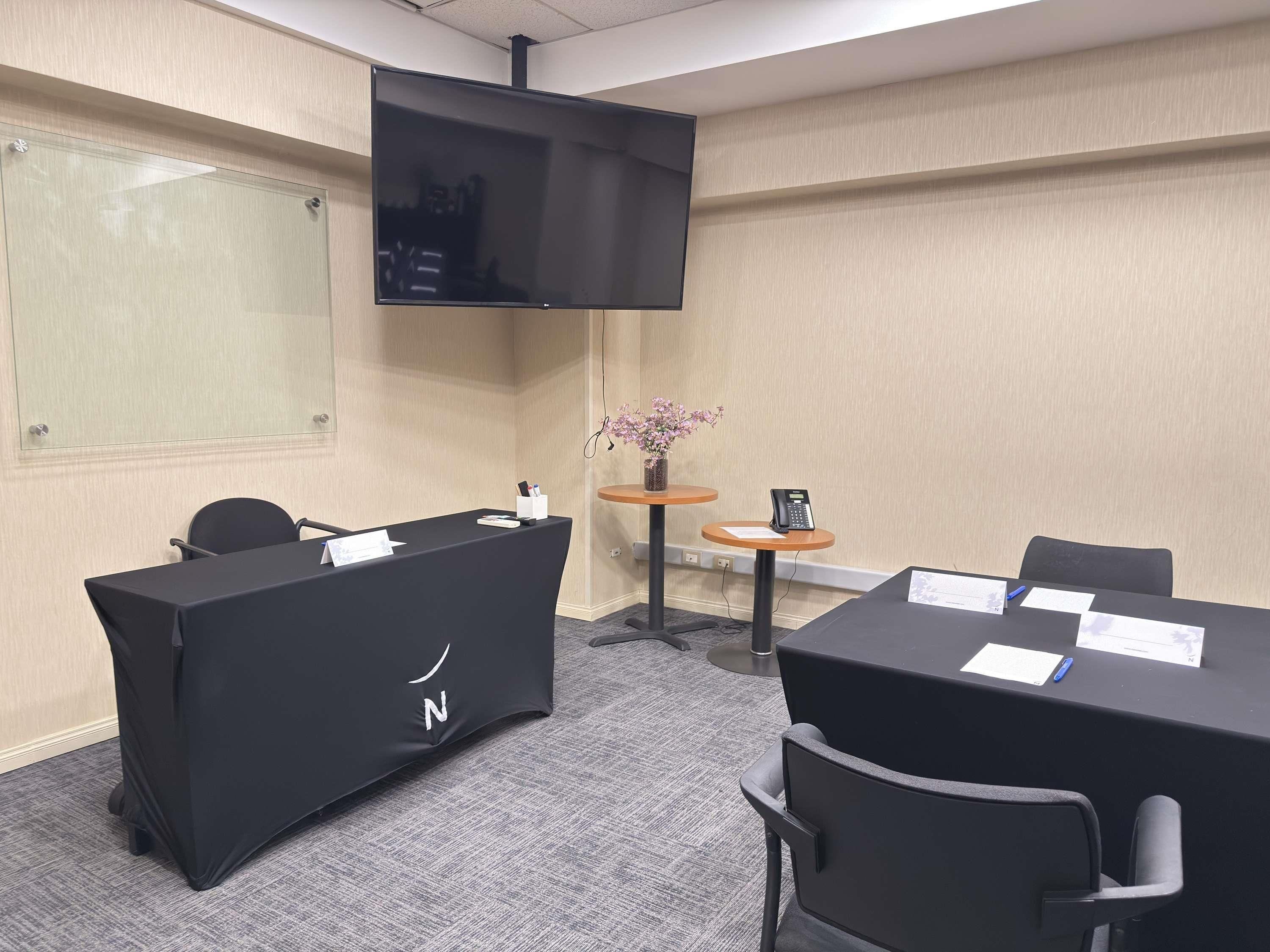 A conference room with a black table, chairs, a TV mounted on the wall, and a small table with a phone and flowers.