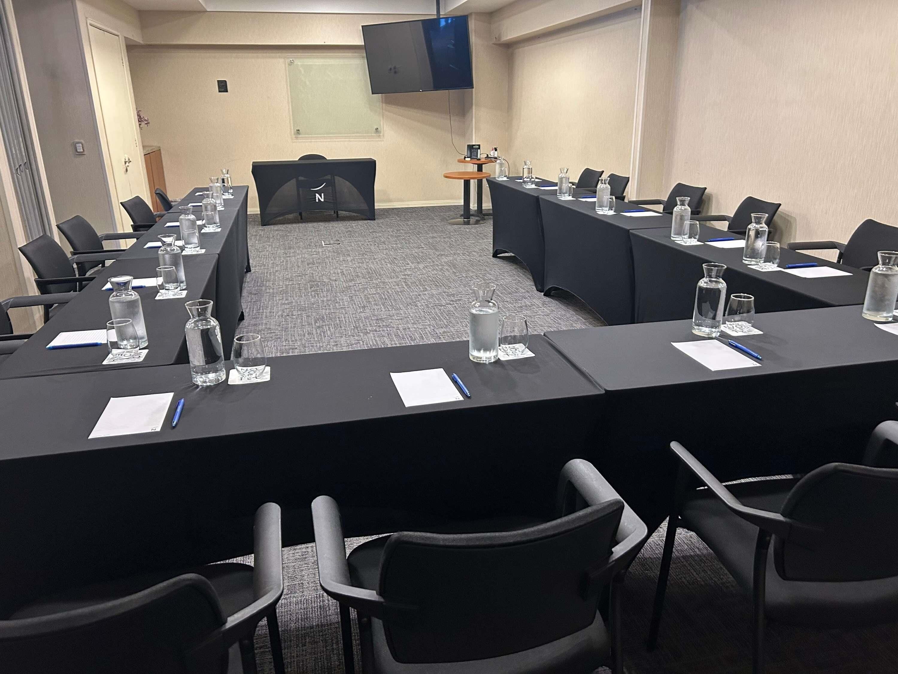 Un salón de conferencias con una larga mesa, sillas, botellas de agua y una pantalla de proyector.