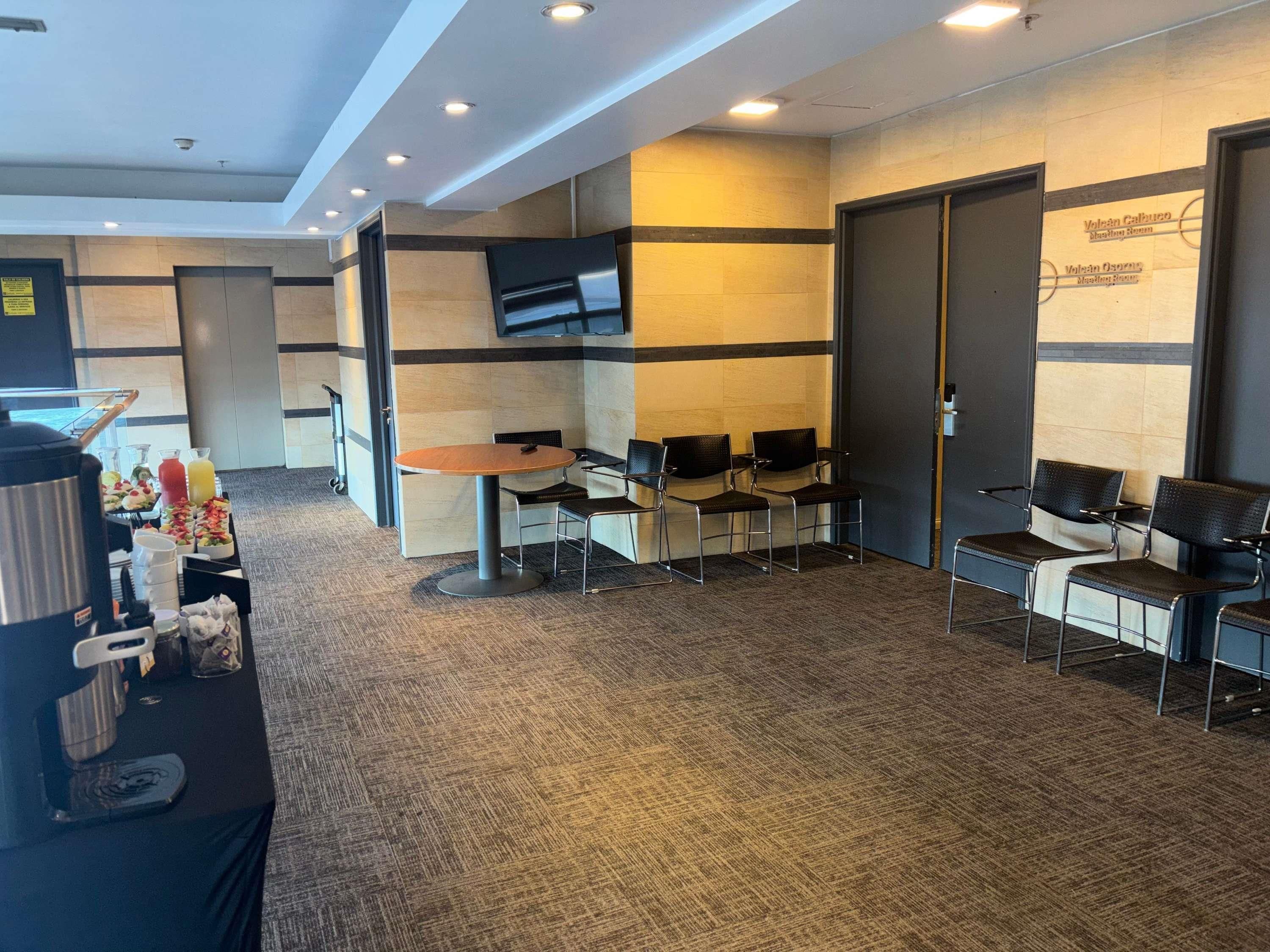 A hotel lobby with a reception desk, a seating area with chairs, a coffee station, and a flat-screen TV mounted on the wall.