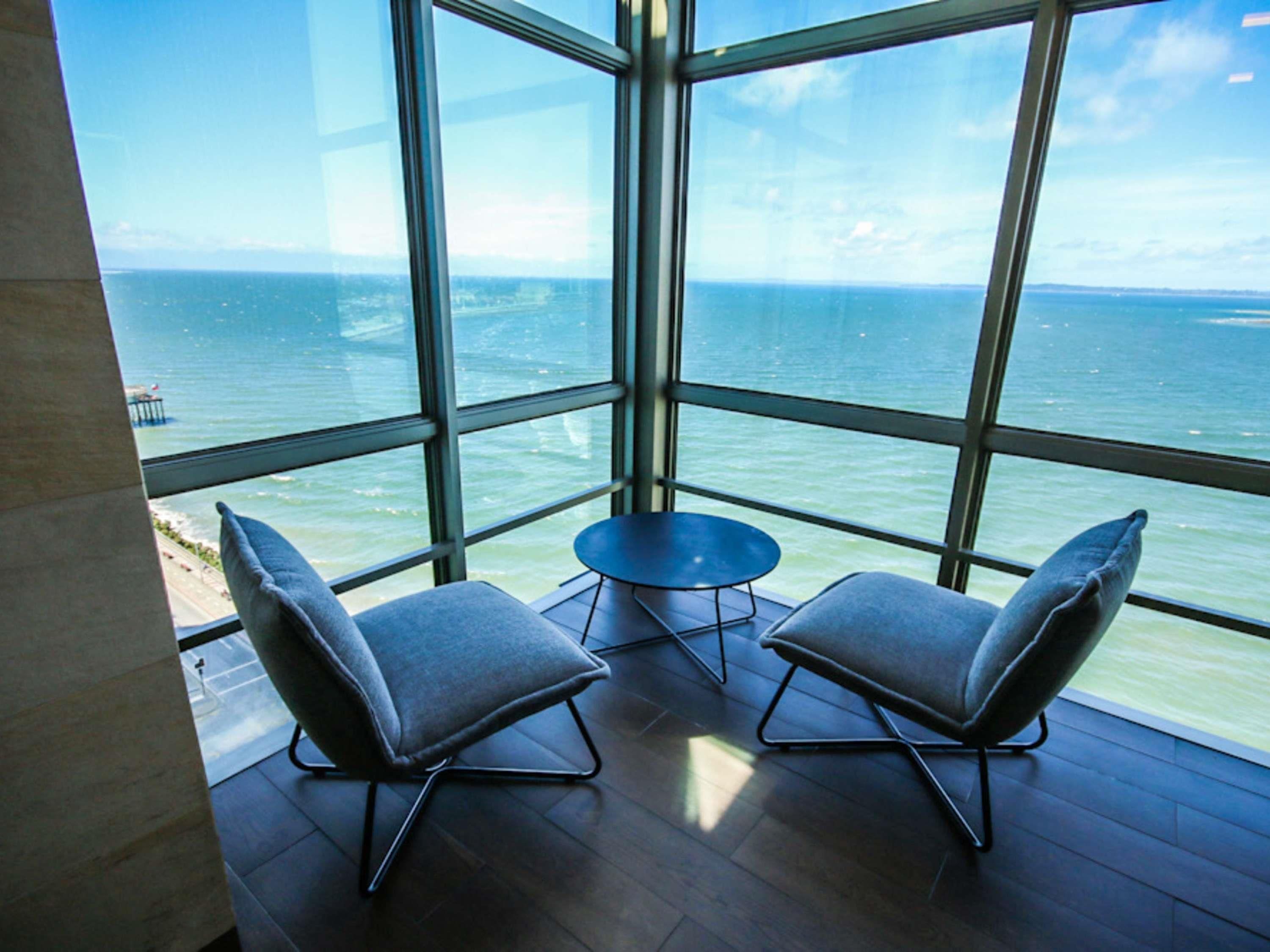 A room with a large window overlooking the sea, two chairs, and a small table.