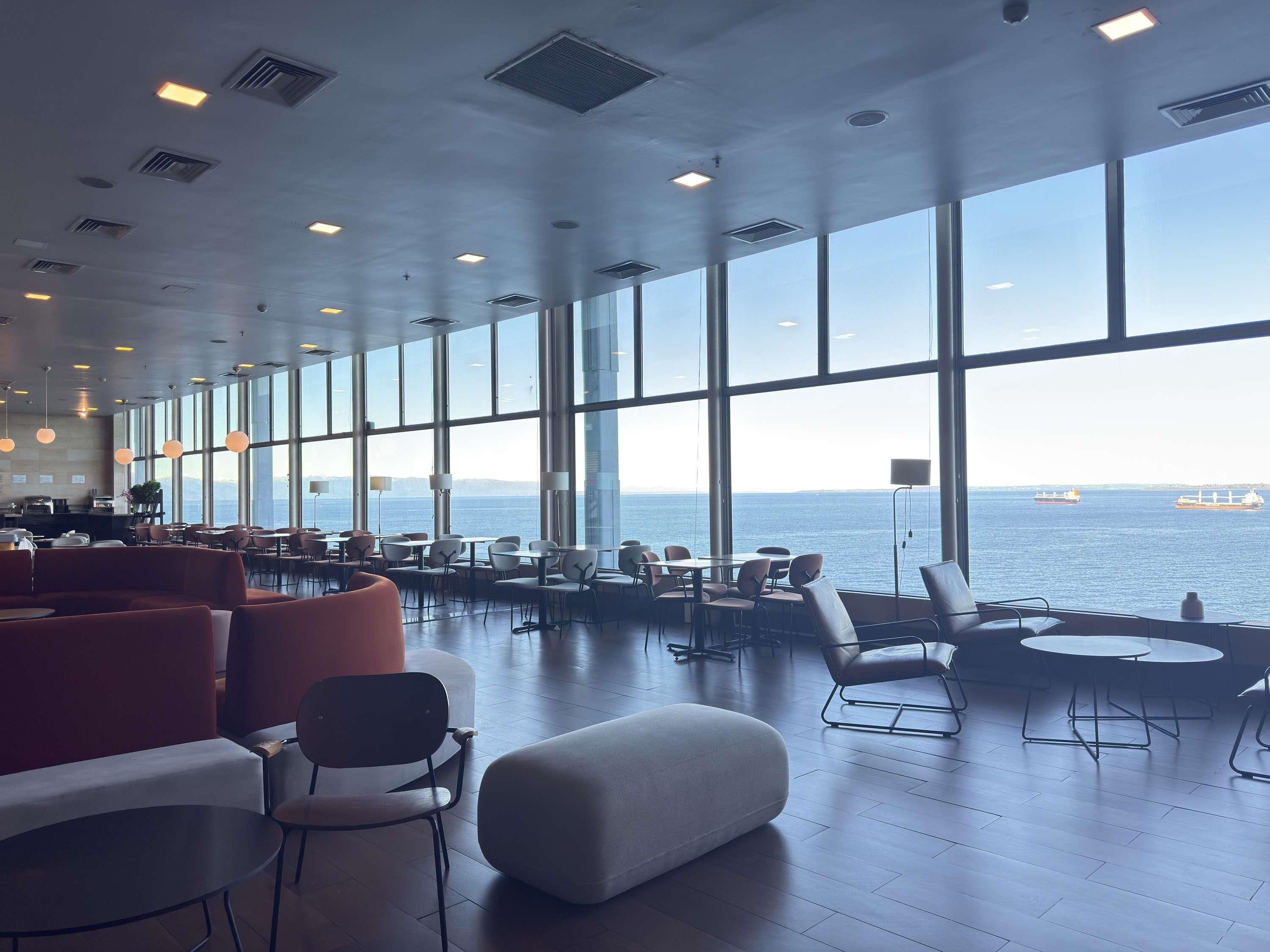 A spacious indoor area with large windows, seating arrangements, and a view of the sea.