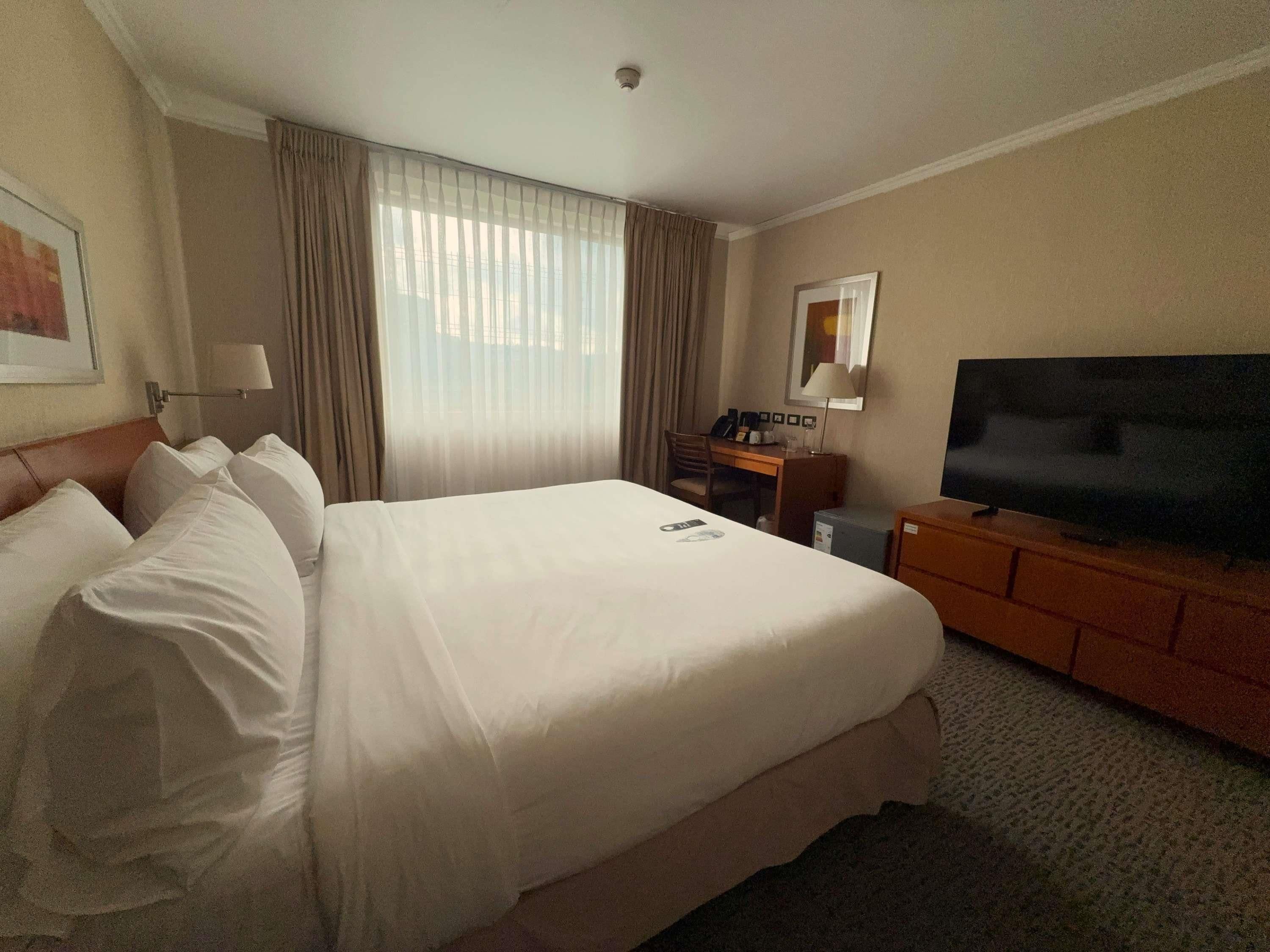Une chambre d’hôtel avec un grand lit, un bureau avec un ordinateur, une télévision et une fenêtre avec des rideaux.