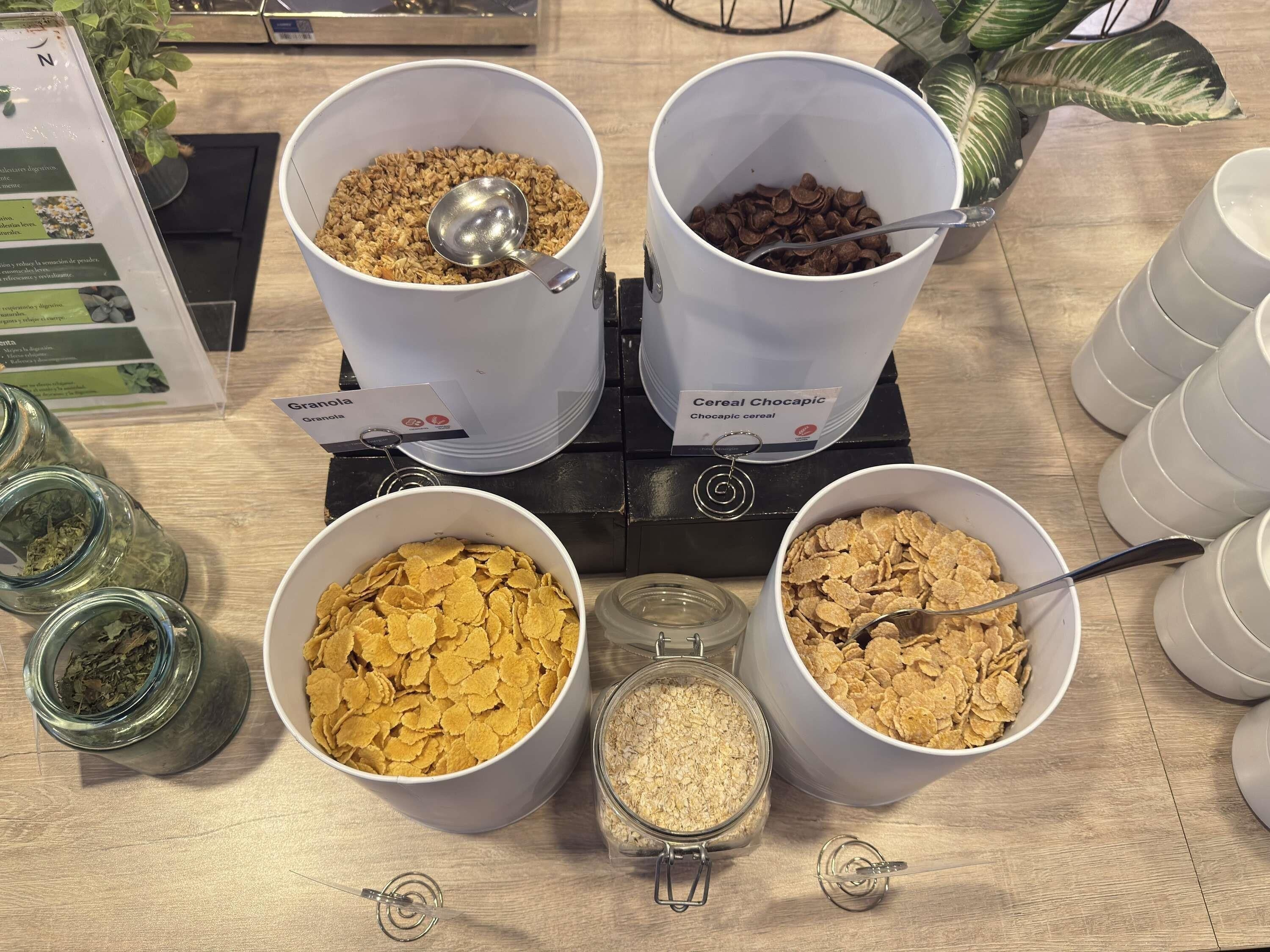 Pöydällä on neljä kulhoa, joissa on eri tyyppisiä muroja, kaksi purkkia kaurahiutaleita sekä kyltti, jossa lukee ”Granola” ja ”Cereal Chocapic”.