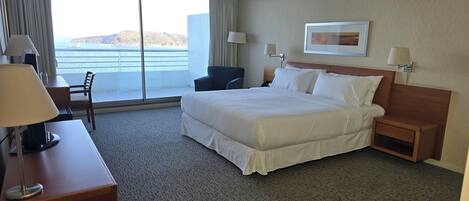 A hotel room with a large bed, a desk with a lamp, a chair, a nightstand, and a view of the sea.