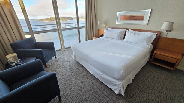 A hotel room with a large bed, a desk, a chair, a nightstand, a lamp, and a view of the sea. - Novotel Puerto Montt (Puerto Montt)