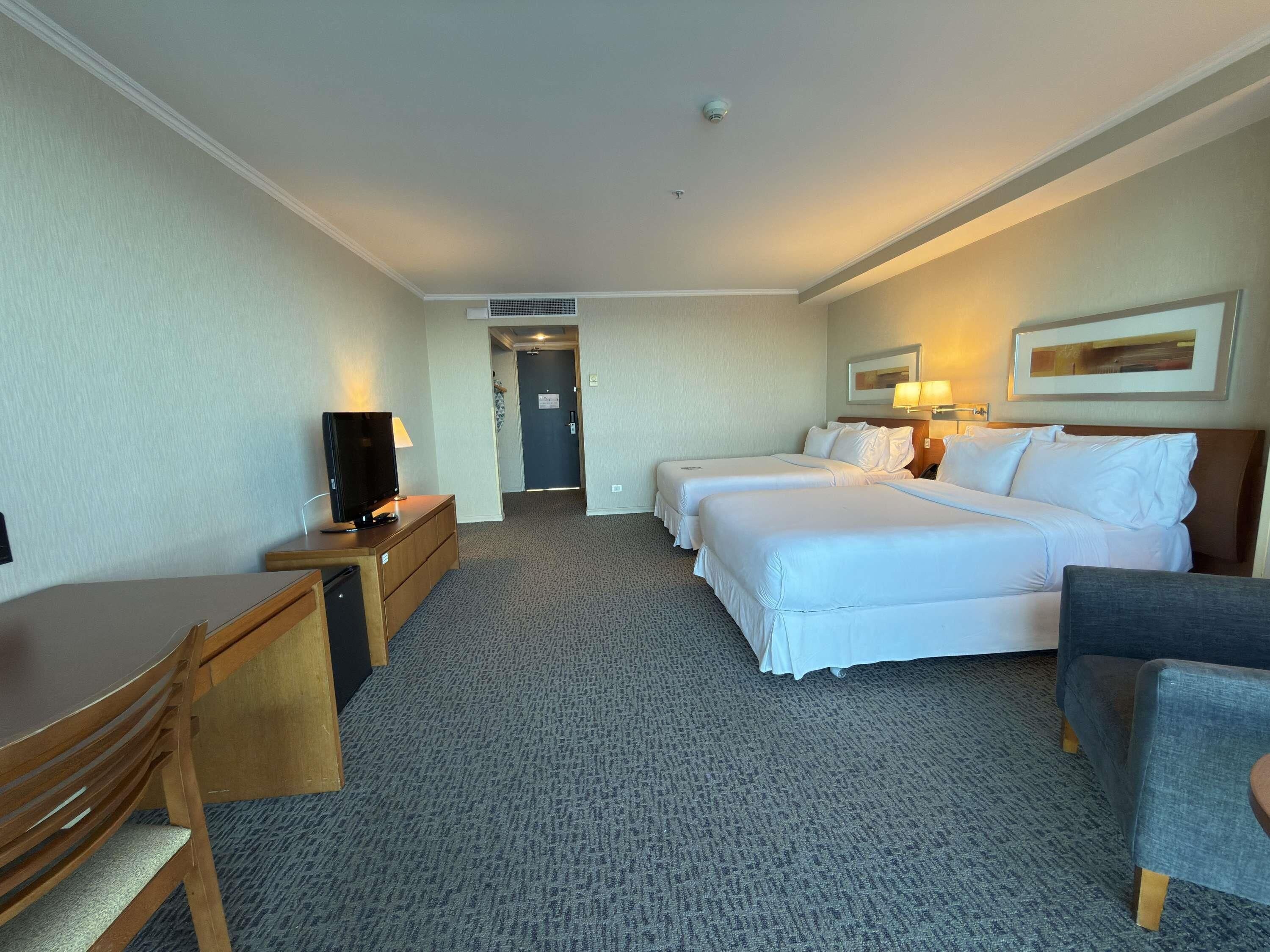 Ein Hotelzimmer mit zwei Betten, einem Schreibtisch mit Fernseher, einem Sessel und Teppichboden.