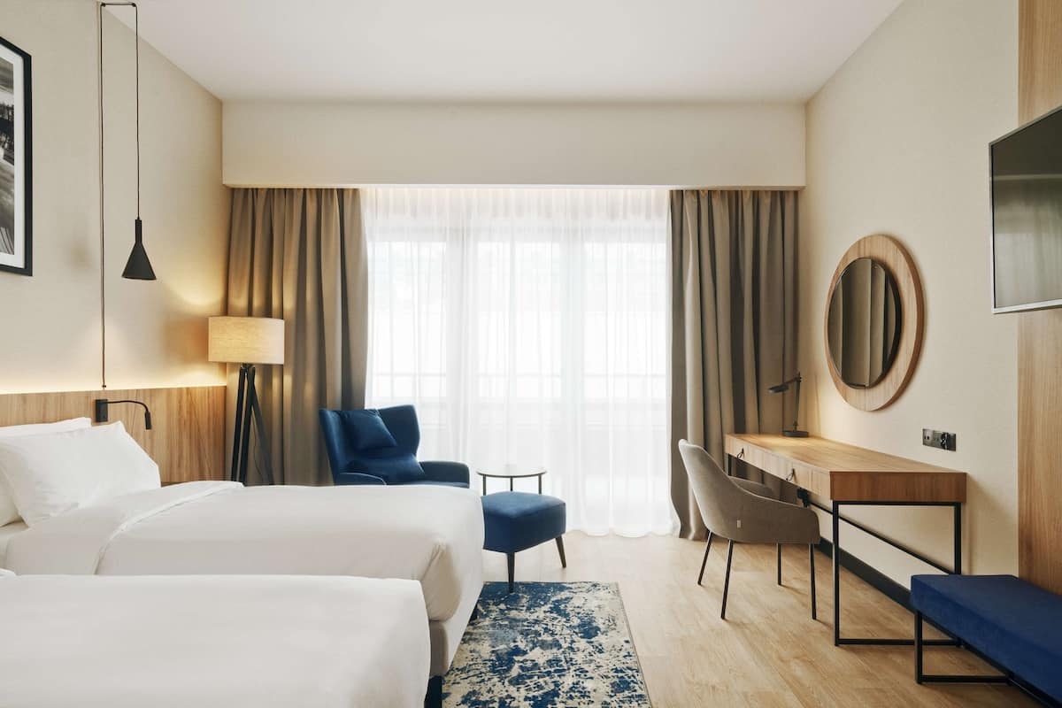 A hotel room with two beds, a desk, a chair, a TV, and a large window with curtains.