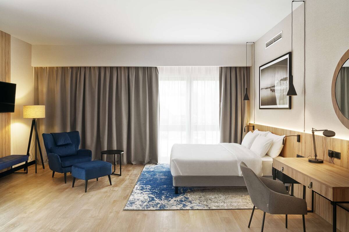 A modern hotel room with a bed, a desk, a chair, a TV, and a large window with curtains.