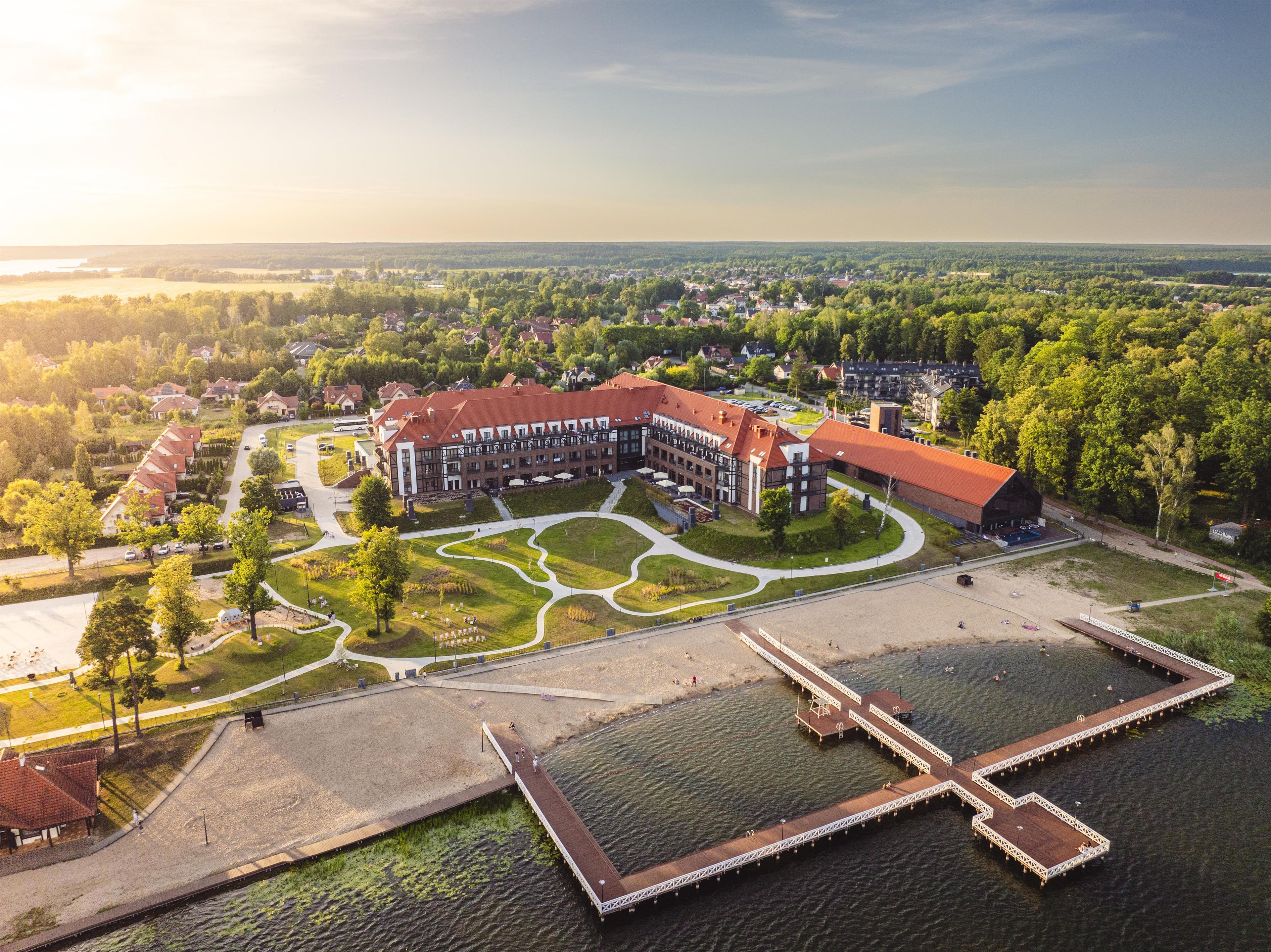 A large building with a red roof, surrounded by a landscaped area with pathways and a dock extending into a body of water.