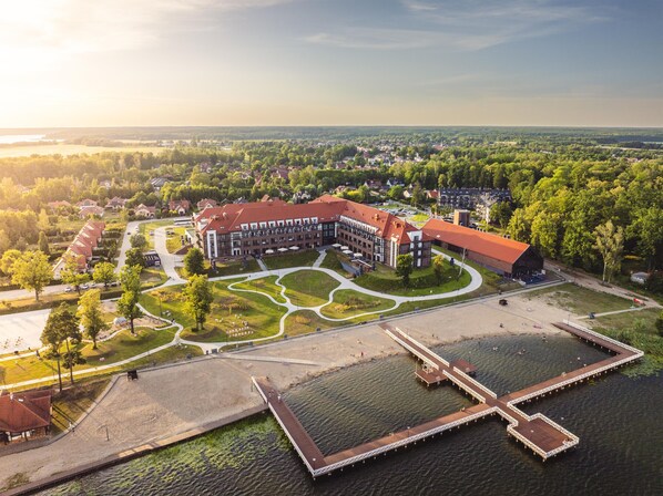 A large building with a red roof, surrounded by a landscaped area with pathways and a dock extending into a body of water.