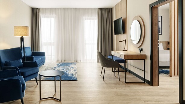 A modern hotel room with a blue sofa, a desk with a mirror, a TV, and a bed in the background.