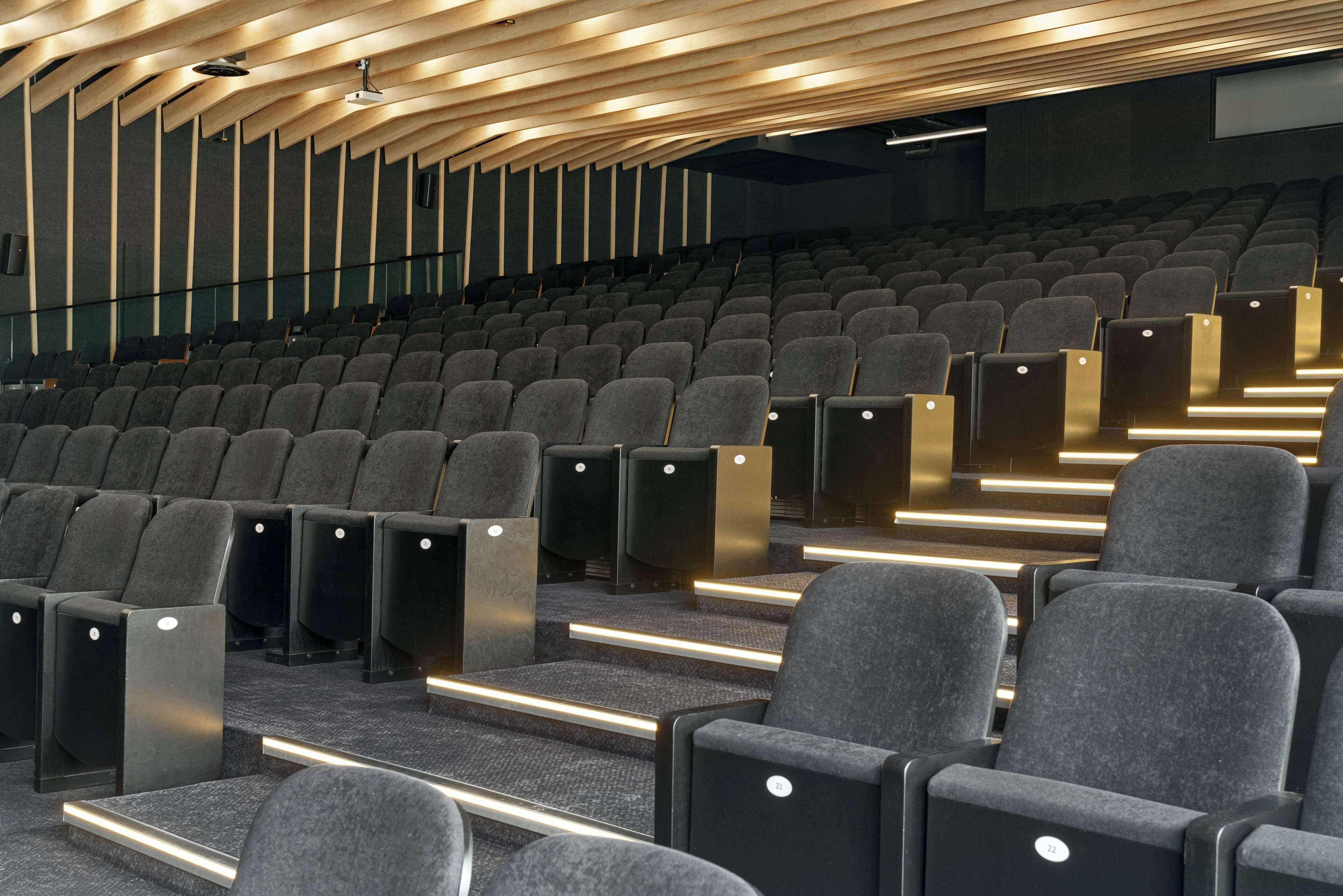 An auditorium with rows of grey upholstered seats and a modern wooden ceiling design.