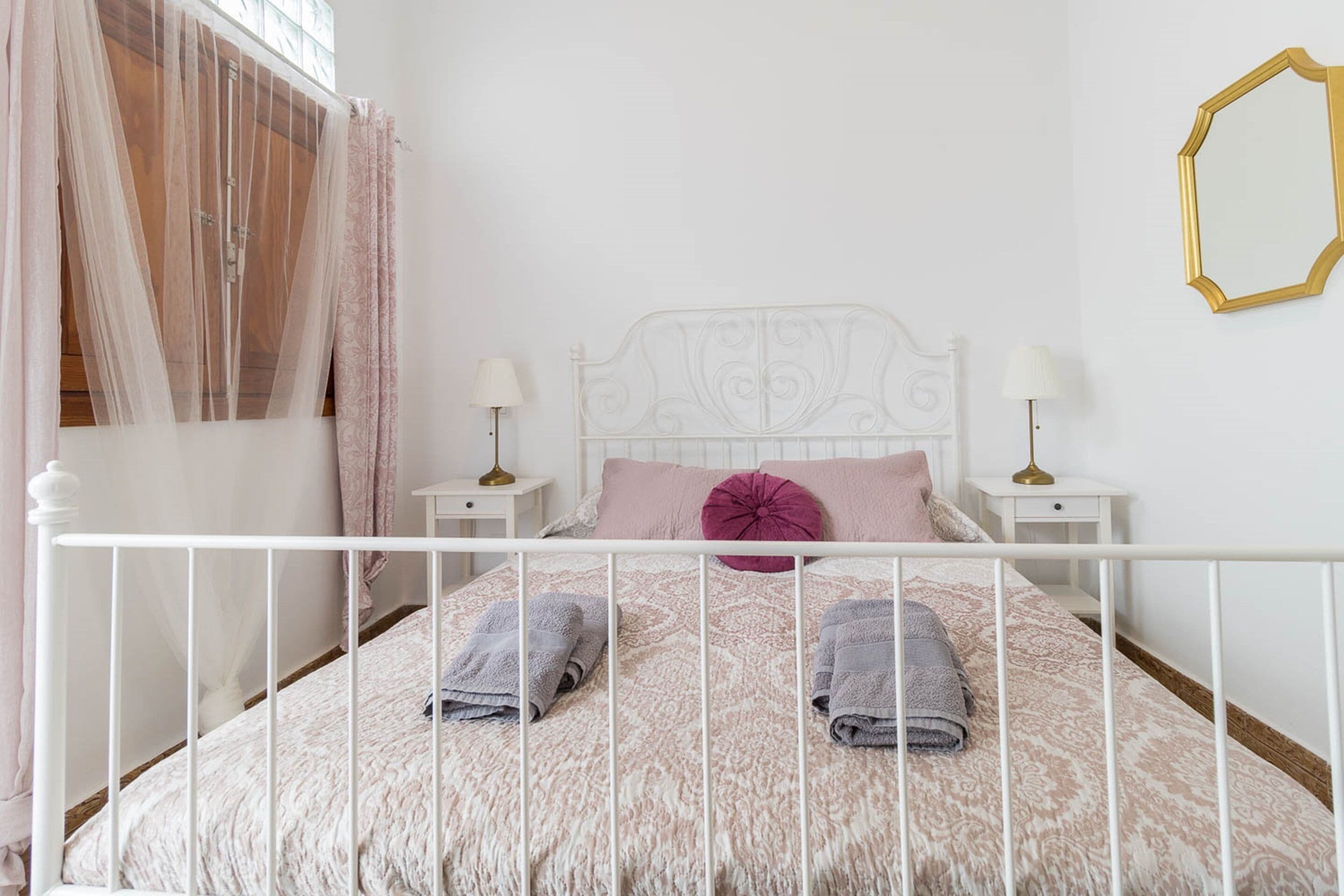A neatly made bed with a white metal frame, two pillows, and folded towels. There are two bedside tables with lamps.