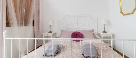 A neatly made bed with a white metal frame, two pillows, and folded towels. There are two bedside tables with lamps.