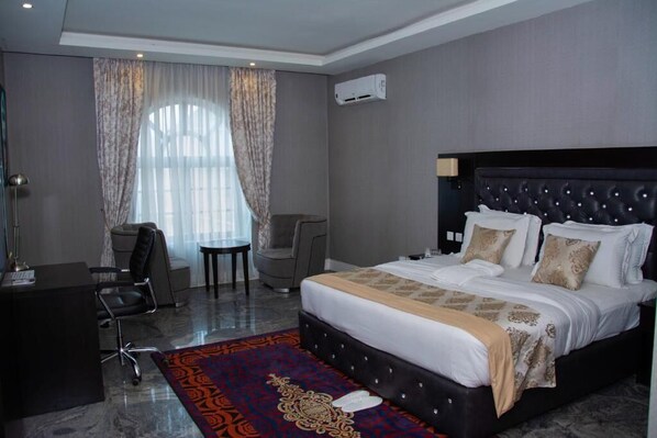 A hotel room with a large bed, a desk, and a chair. There is an air conditioning unit on the wall and a window with curtains.