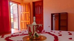Une chambre avec un lit recouvert de pétales de rose, un vase décoratif et une vue sur l’extérieur par une porte ouverte.