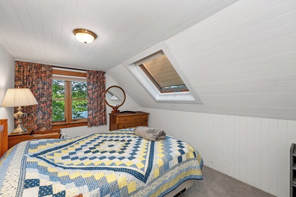 A bedroom with a sloped ceiling, a skylight, a bed with a patterned quilt, a wooden dresser, a lamp, and a window with curtains.