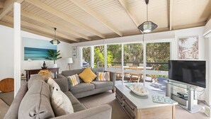 A spacious living area with a grey sofa, a coffee table with decorative items, a TV, and a large window overlooking a deck with outdoor furniture. - Amongst The Ti-Trees - Rye (Rye)