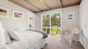A bedroom with a bed, a wicker chair, and a large window with a view of trees. - Amongst The Ti-Trees - Rye (Rye)