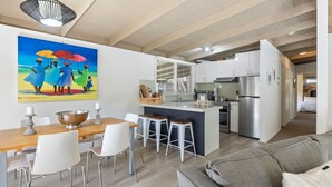 A modern kitchen with white cabinets, a stainless steel refrigerator, and a dining area with a wooden table and white chairs. - Amongst The Ti-Trees - Rye (Rye)