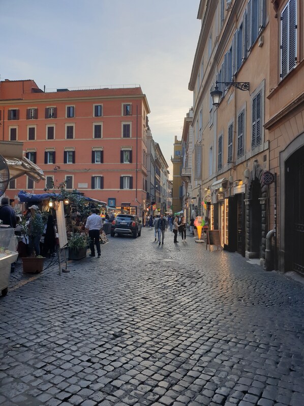 A cobblestone street with buildings on either side, a market stall with plants, and a few pedestrians. - Brand-new, elegant, and cozy apartment in the heart of Rome. For couples or families: Wi-Fi, air conditioning (Roma)