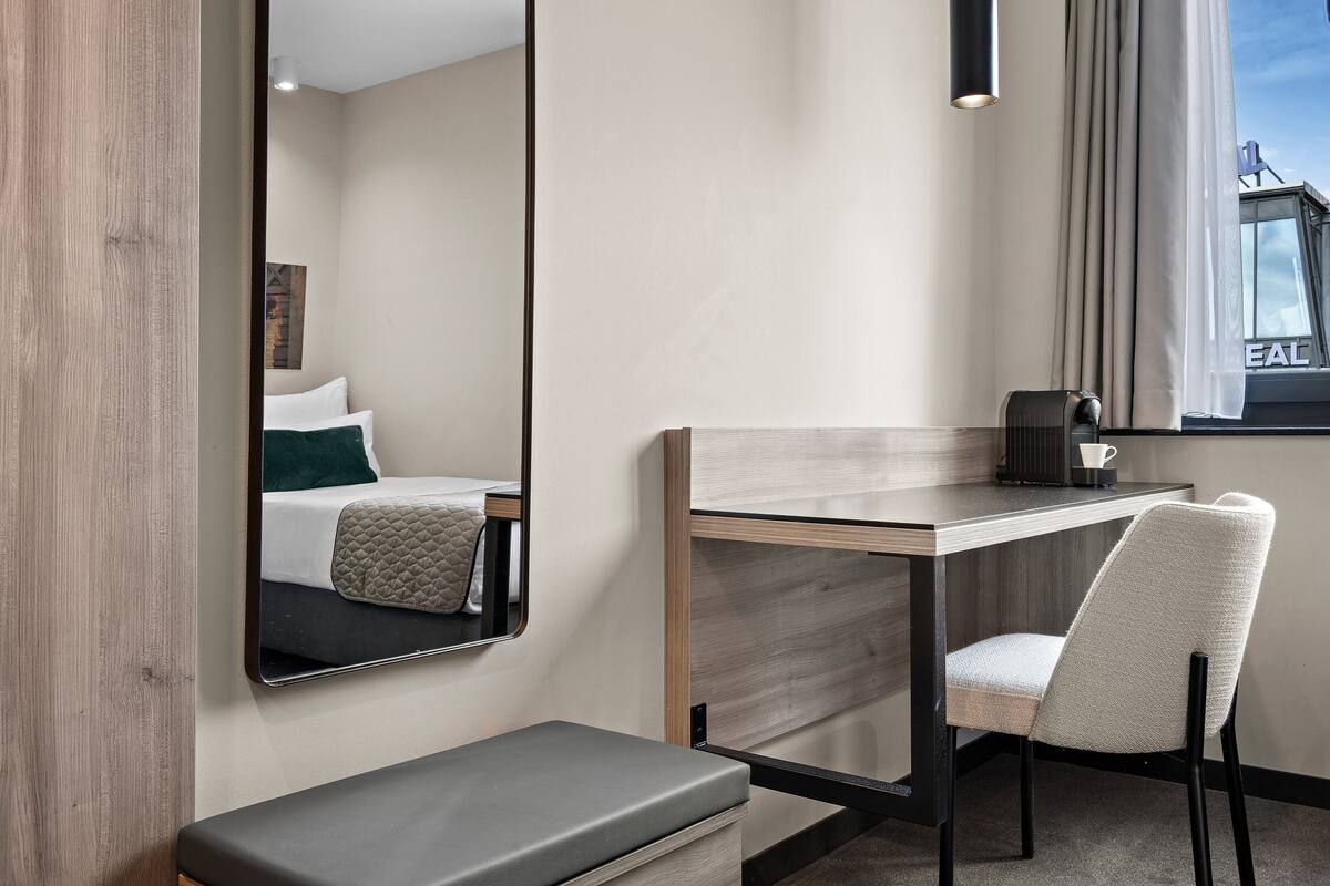 A hotel room with a bed, a desk, a chair, a bench, and a large mirror.